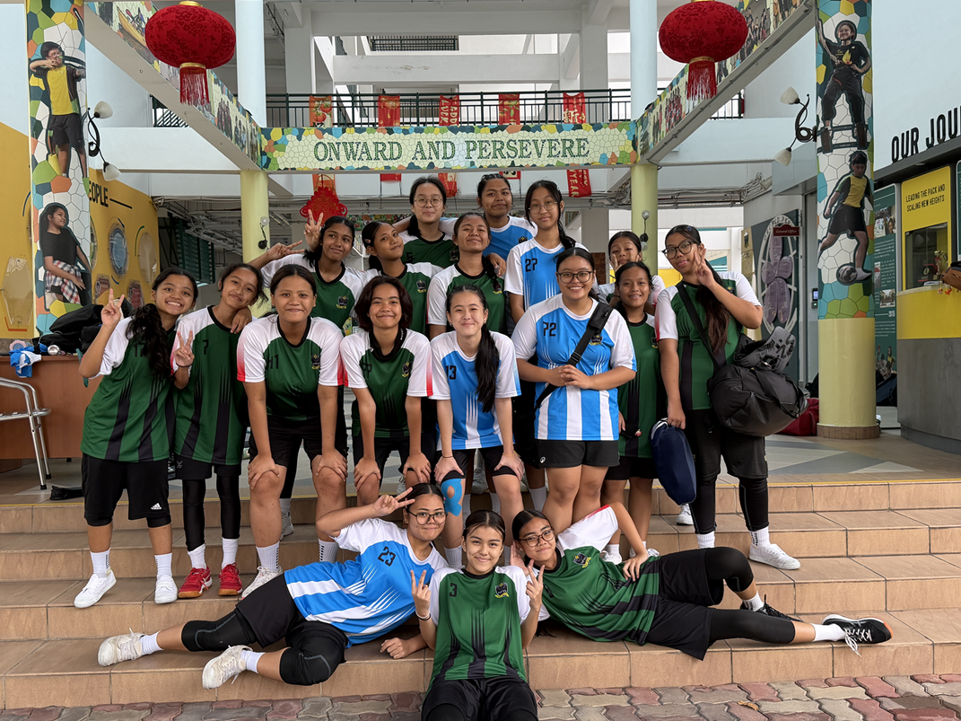 A girls' floorball team smiling and posing together on school steps decorated with red Chinese New Year lanterns. The players are wearing a mix of green-and-black and blue-and-white sports jerseys, positioned under a sign reading "ONWARD AND PERSEVERE".